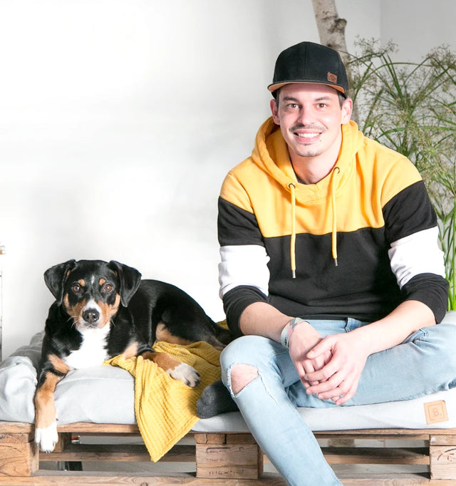 BUDDY Hundezuebhör Gründer Manuel sitzt mit seiner Appenzellersennenhündin Momo auf einem grauen BUDDY. Hundebett. BUDDY Hundezuebhör Gründer Manuel sitzt mit seiner Appenzellersennenhündin Momo auf einem grauen BUDDY. Hundebett.